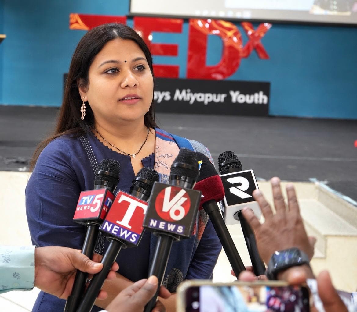 Pushpa Valli speaking with reporters in front of a TEDx event backdrop.