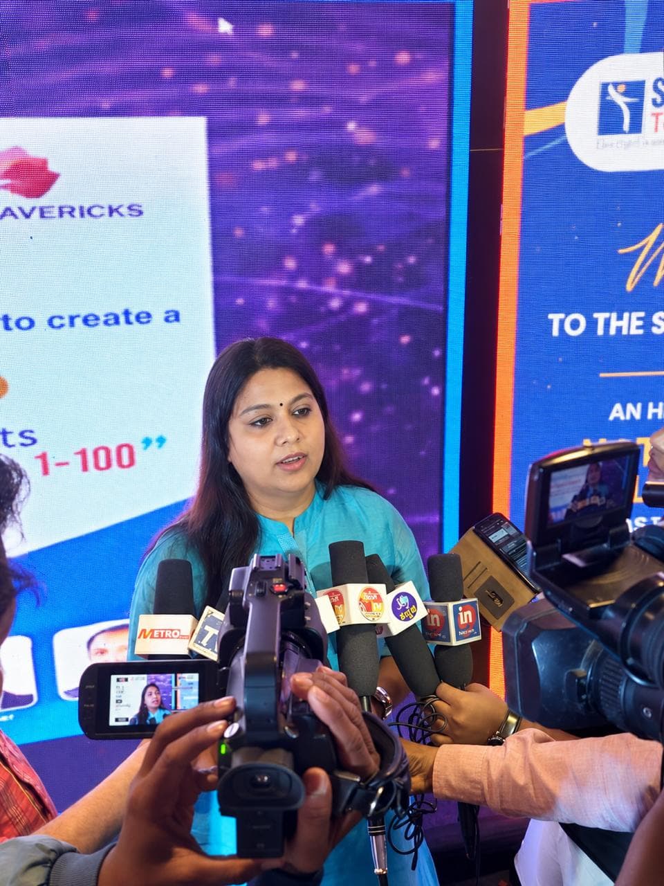 Pushpa Valli speaking with journalists, surrounded by microphones and cameras at a Mavericks event.