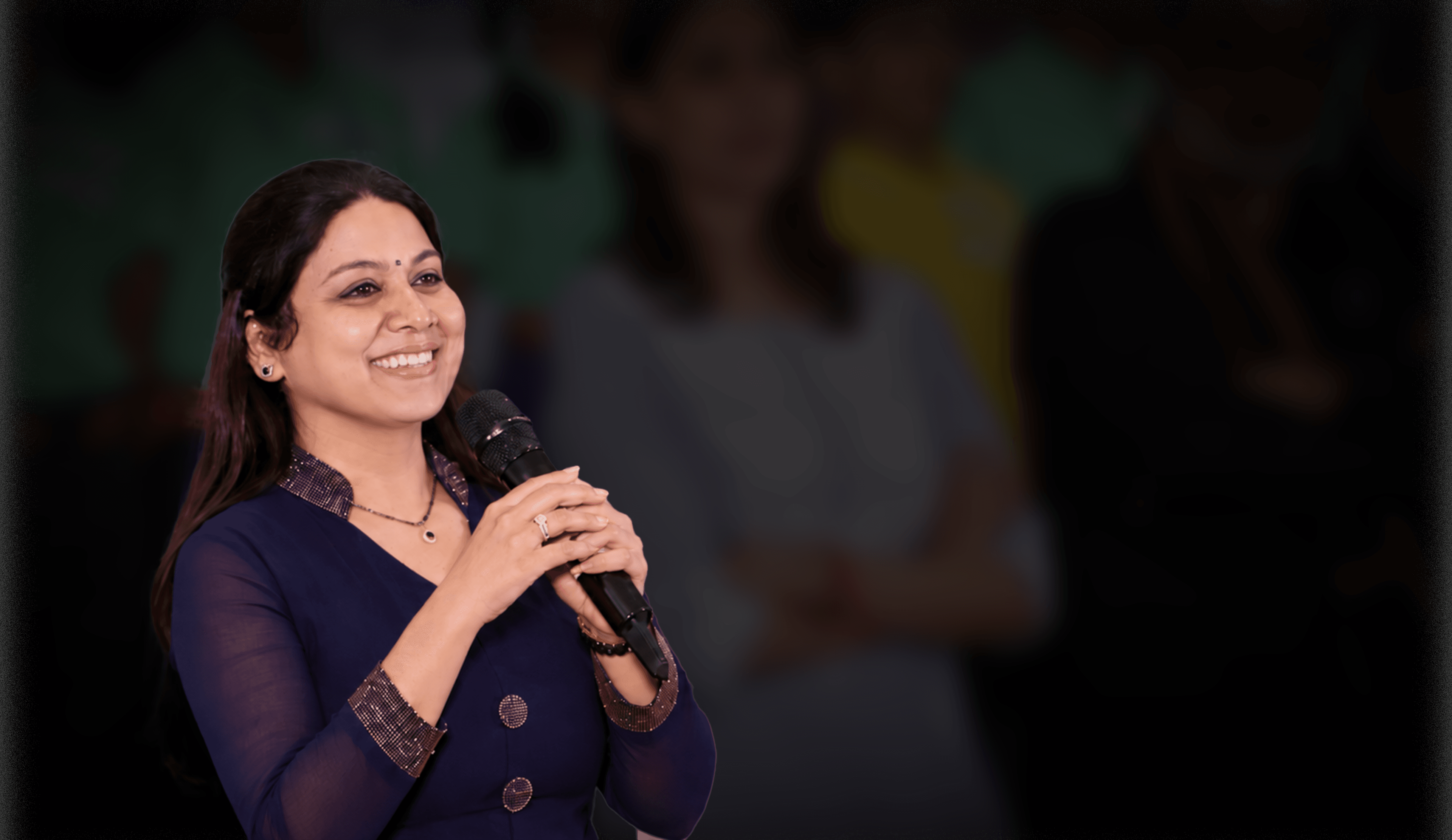 Pushpa Valli smiling and speaking into a microphone against a softly blurred event backdrop.