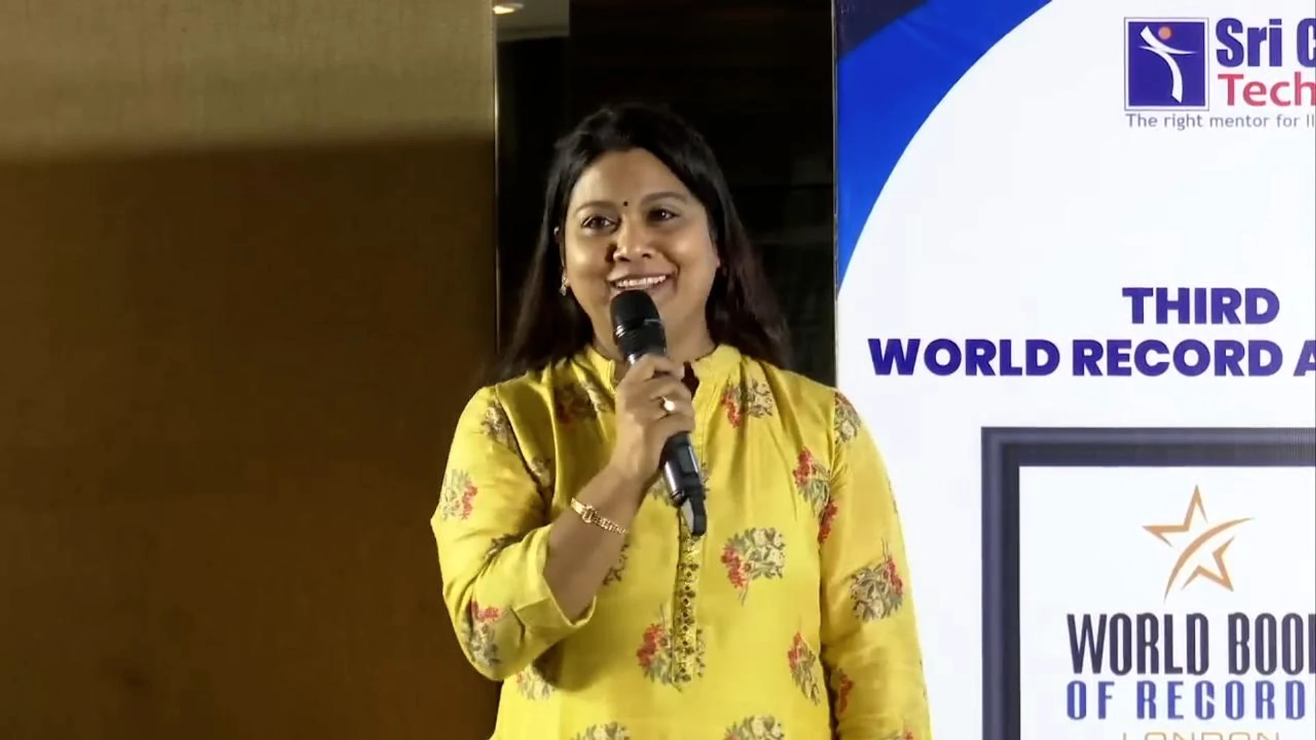 Pushpa Valli speaking into a microphone beside a world record event banner.